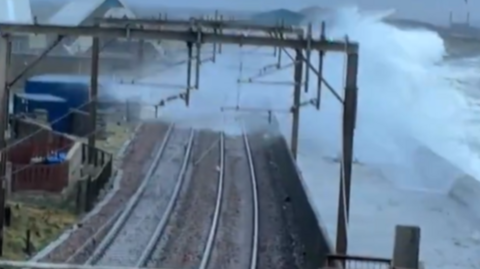 Large waves crash over a sea wall next to railway tracks, with water spraying onto the line. Overhead power lines and poles run along the track under a cloudy, stormy sky.
