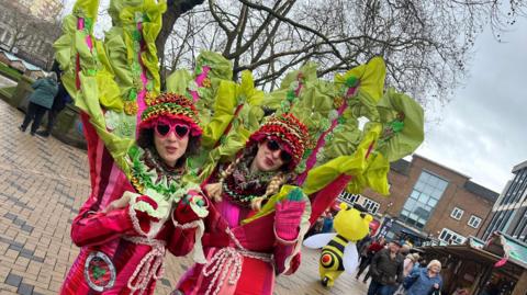 Two women dressed in rhubarb stalk costumes in shades of pink and red, with large green leaf‑like structures extending high above their heads. They are standing outside in Wakefield town centre, with paved pedestrian walkways and wooden stalls behind them.