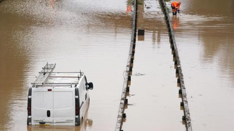A white van is stranded in floodwater on the A189 Spine Road near Blyth, Northumberland. The water comes up to the the top of the vehicle's number plate. A worker in an orange high-vis uniform is crouched down further up the road.