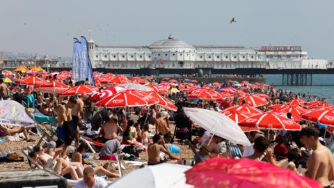 Hundreds of people under red umbrellas on Brighton beach with the Palace Pier in the background
