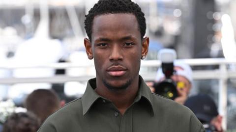 Micheal Ward, surrounded by photographers and wearing a dark green shirt and earring, looks straight into the camera