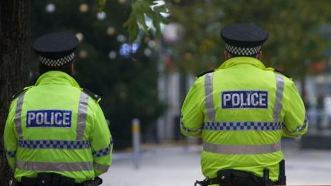 Two police officers stand with their backs to the camera. Their uniforms are florescent green jackets with the word POLICE in white block capitals on a blue background. Liverpool city centre's Lord Street can be seen in the background, out of focus