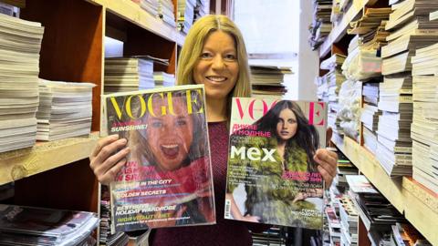 A blonde woman smiling at the camera standing in the middle of two shelves that reach the ceiling and are full of magazines. She's holding two vintage Vogue magazines in front of her, one in each hand. The magazine on the right shows a mid shot of a brunette model looking into the camera with her hair blowing in the wind and she's wearing a green fluffy coat. The cover on the left shows a close up portrait of a blonde model smiling wide looking into the camera holding a quarter of watermelon.