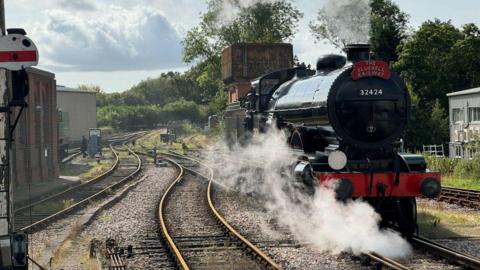 A black locomotive runs along railway tracks as a jet of steam comes out the top