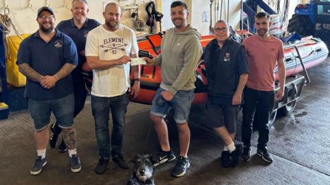 Curtis Johnson is wearing a grey hooded top, denim jean shorts and black trainers, and holding the lead to Rex, a black terrier-type dog. He is handing a cheque to one of the five male members of crew, who are all casually dressed. Three of them are wearing branded RNLI polo shirts. They are standing in the lifeboat station in front of the inshore lifeboat and everyone is smiling.