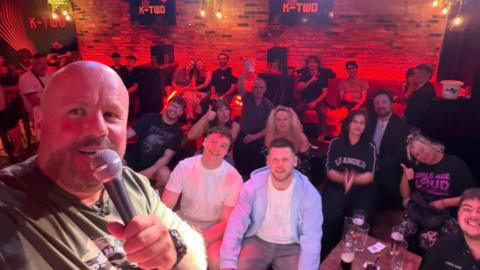 Selfie style group photo with Russ Lee on the far left. He is wearing a khaki t-shirt and is holding a microphone. 