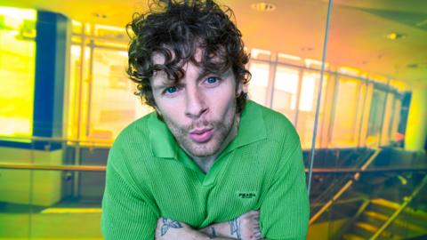 A man with curly hair and visible tattoos on his arms is wearing a green collared Prada shirt. They are indoors in a space with glass walls and warm yellowy/orange lighting. The man is leaning slightly forward, facing the camera and is pouting his lips.