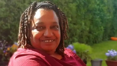 Marcia Grant smiles at the camera. She has shoulder length black hair and is wearing a red top. She is sitting in a garden. 
