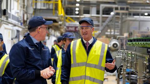 Peter Kyle being led around a glass factor in Cheshire - he is gesturing with his left hand, while standing among the machinery - he is wearing a baseball cap and a high visibility jacket