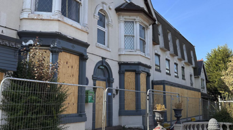 A hotel site with at least half a dozen silver barriers in front of it. Windows and a door on the ground floor are boarded up on this photo that was taken outside.