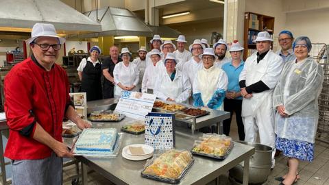 Laurence Wells in the foreground holding a cake while his colleagues pose with food gifts and cards in the hospital kitchen