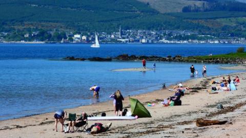 People sit on the sand by the waterside on a sunny day. A town can be seen in the distance across the water