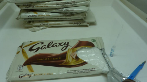 The wrapping of a Galaxy chocolate bar has been ripped open to reveal a bag of white powder. More chocolate bars are in a pile behind it and there is a scalpel and a drug test swab next to it.