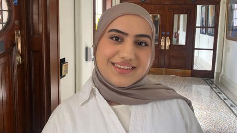 A woman standing in a corridor, she is wearing a brown hijab with a white shirt.