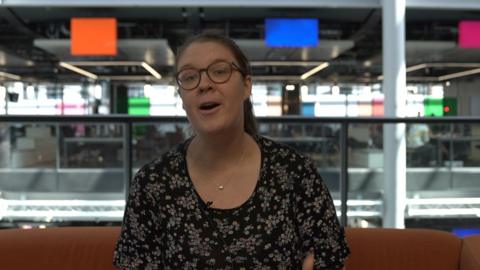 Catriona Aitken - who has glasses and tied-back brown hair and is wearing a black floral dress - sat on an orange sofa, with the colourful panels of the 鶹ҳ Wales office behind her