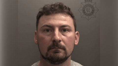 A police custody photo of Peter Coope. He has short brown hair and a short brown mustache and beard. He is wearing a grey t-shirt and is looking at the camera. The wall behind him is grey.