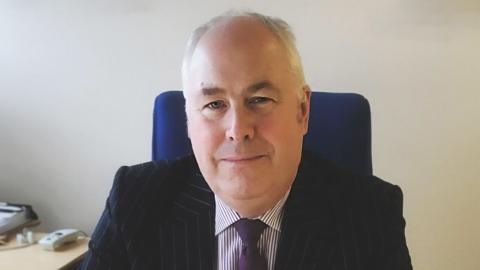 Malcolm Burr - a man with grey hair pictured sitting at his desk smiling wearing a pinstripe suit and purple tie