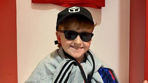 A young boy sits in a football changing room smiling at the camera. He has both thumbs raised up. He wears a black cap and sunglasses and has a grey jumper tied around his neck.