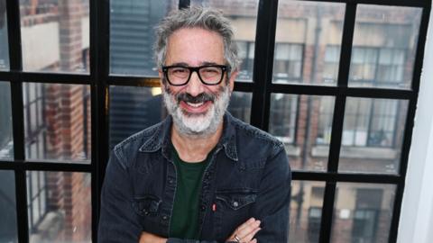 A man with black rimmed glasses and white and dark hair, with a beard, smiles at the camera, with his arms crossed against his chest, standing in front of a black framed window, and brick building 