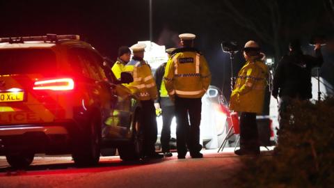 Four police officers stand beside a patrol car in Grace Road, Crawley, with the scene illuminated by a spotlight.