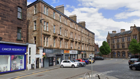 Streetscape showing a row of sandstone tenement buildings with ground-floor shops, including a Cancer Research UK store, parked cars along a cobbled street, bicycle racks in the foreground, and a historic stone building in the background under a partly cloudy sky
