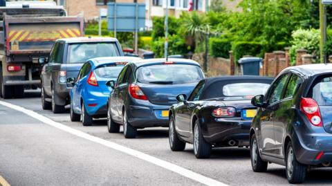 A queue of cars form traffic next to a bus lane on the left and a row of low walls and gates to front gardens with shrubs in leaf on the right.