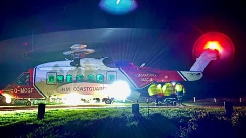 The picture shows a Coastguard rescue helicopter on the ground at night with its lights blazing so brightly that they spill across the grass around it. The rotors are spinning, creating a blurred circular halo above the aircraft. The helicopter’s red and white body stands out against the darkness, and the tail light glows with a bright red ring.