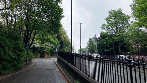 Footpath next to the eastbound side of a dual carriageway. There are a lot of cars on the westbound side but the nearest carriageway has no cars on it. There are black railings between the path and the road. On the other side of the path - and also next to the far carriageway - there are lots of trees and bushes.