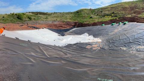A large black and white tarpaulin on a wide circular area of ground in the countryside. Grass can be seen in the distance and red earth at the edge of a sunken area that has been excavated. It is a sunny day.