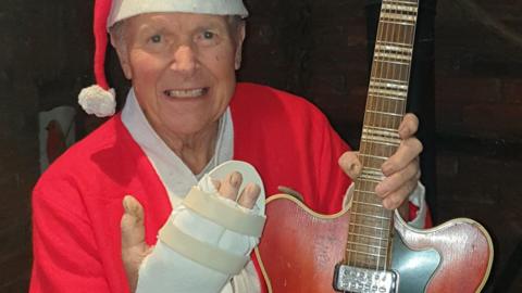 Mr Dawson wears a santa outfit and holds up his bandaged hand.  In his other, he holds his guitar