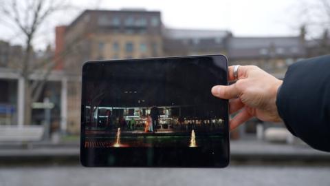 An iPad showing a scene in Virdee in the centre of Bradford with the actual setting blurred behind it.