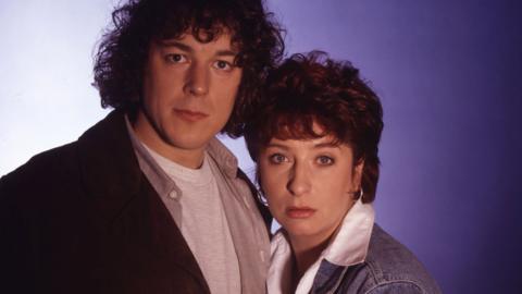This image shows two adults posed close together against a plain, softly lit background that fades from light grey on the left to a bluish tone on the right.
