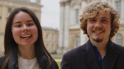 A man and a woman smile at the camera. The man has short curly blond hair and the woman has long dark hair. They are both wearing black jackets. The man has a blue shirt under his jacket while she has a white top on underneath. University buildings are behind them.