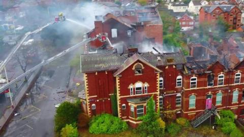 Aerial image of firefighters, standing on a cherrypicker platform, spraying water down on to the roof of a four-storey, redbrick building below.