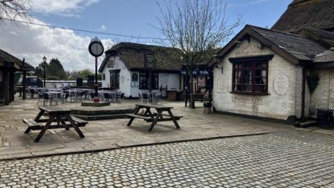 Picnic tables and a large clock are situated outside a thatched building.