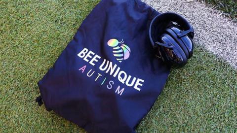 A dark blue drawstring bag lying on a football pitch with the words "Bee Unique Autism" on the bag and ear defenders resting on top