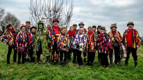 Members of Sompting Village Morris posing for a photograph while standing in an open green field. They are dressed in colourful garbs and black hats with flowers on them, accompanied by a man with a painted green face and green trench coat.