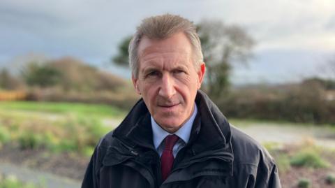 Dan Jarvis is looking serious and at the camera. There is a garden out of focus behind him. He has short grey hair and blue eyes. He is wearing a black waterproof jacket and a blue shirt with a burgundy tie.