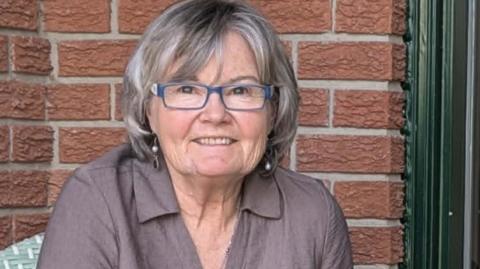 An image of Elizabeth Doran. She is smiling and has a short grey bob with bangs, blue-framed eyeglasses and dangling earrings. She is wearing a collared linen grey-mauve shirt, and behind her is a brown brick backdrop. 