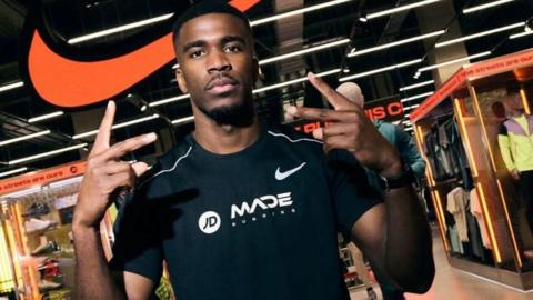 Hermen Dange hold up his arms while he poses in a black t-shirt outside a shop