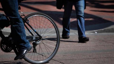 A generic image of a person riding a bicycle along a road while another person in the background walks across the road.