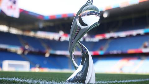 The Women's Euro 2025 trophy standing on a football pitch. The design is modern and fluid—two sweeping arms curve upward to cradle a spherical element at the top, creating a sense of movement and elegance. The metallic surface catches the light, giving the whole trophy a polished, prestigious aura.

In the background, we see rows of blue stadium seats