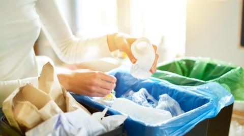 A woman wearing a white long sleeve top placing a plastic bottle in a recycling bin.
