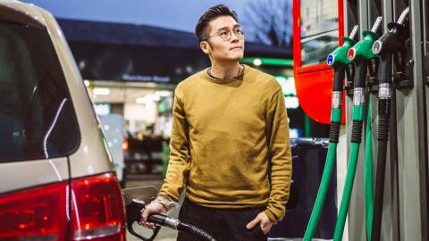 Adult man refuelling his car at a petrol station during evening hours, pausing thoughtfully beside fuel pumps. He is wearing a light brown jumper. 