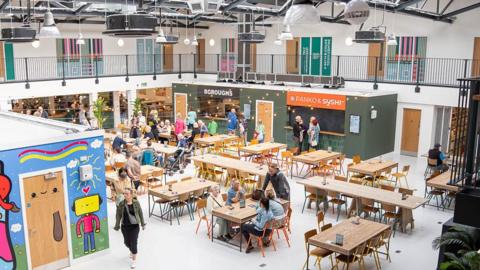 Inside the market, a large space, with painted white walls and flooring. On the lower floor, are central tables full of people with food shops surrounding them. On the first floor, railings, and behind them, a series of doors