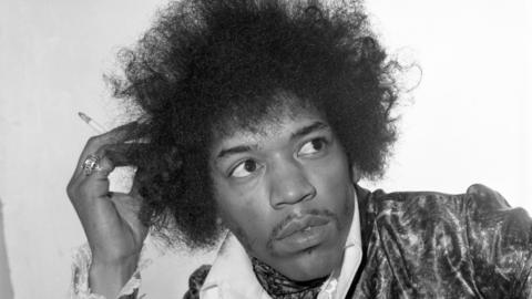 A black and white photo of rock star Jimi Hendrix. He is looking to the right of the shot with a cigarette in his right hand which his head is resting on. He is wearing a collared shirt and a velvet-looking jacket.