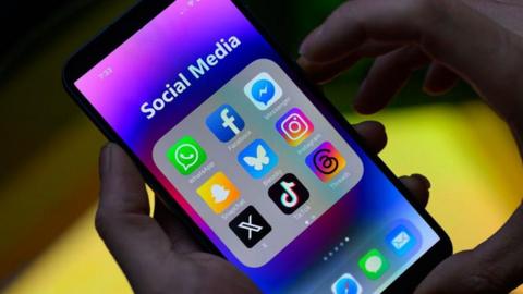 Stock image of different social media apps on the screen of a phone. A person's hands can be seen holding the phone with a dark background. 