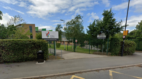 A school building surrounded by trees and hedges. It is a sunny day.