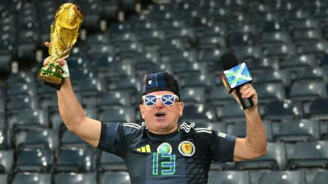 A man clad in Scotland gear holds a replica gold World Cup trophy above his head. He is wearing a tartan hat, Scotland strip and sunglasses with saltires on the lenses. He is standing in front of an empty blue stand. 
