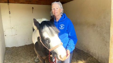 Karen Thompson, who has white hair and is wearing a blue fleece top, stands next to a black and white pony